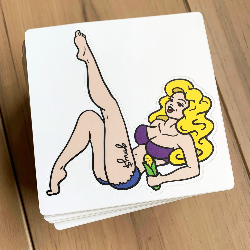 A sticker of a blonde woman on her back, with one foot playfully in the air. She holds an ear of corn, and has a thigh tattoo that reads 